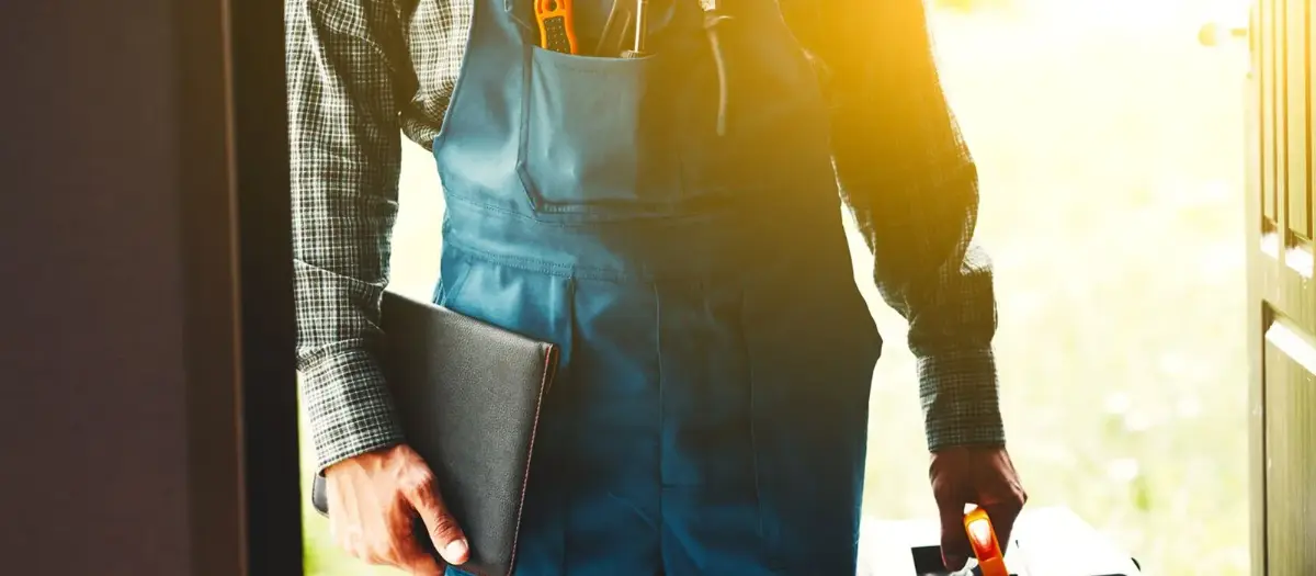 Eine Person in blauer Arbeitskleidung hält einen Laptop in der Hand. Heller Hintergrund.