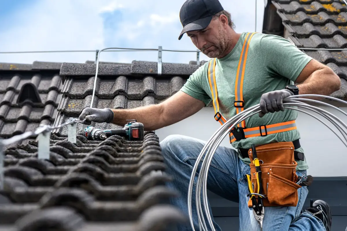 Ein Handwerker auf einem Dach, der mit Werkzeugen Kabel verlegt. Sonniger Himmel.